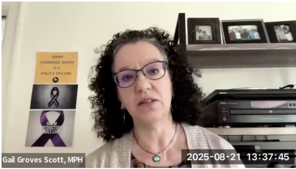 A woman with brown curly hair and glasses is seated in an office and being recorded on a zoom call